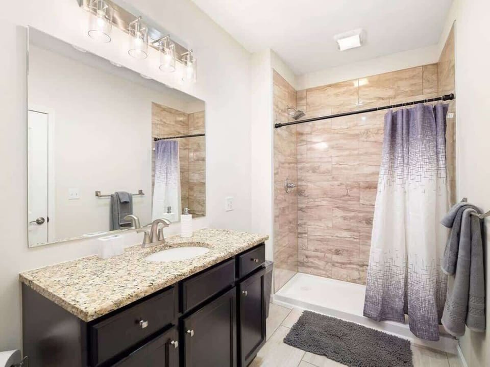 Bright bathroom featuring a granite-topped vanity with dark wood cabinets, frameless mirror, and modern lighting. Spacious tiled shower with sleek fixtures and plush mat—your daily oasis! ✨