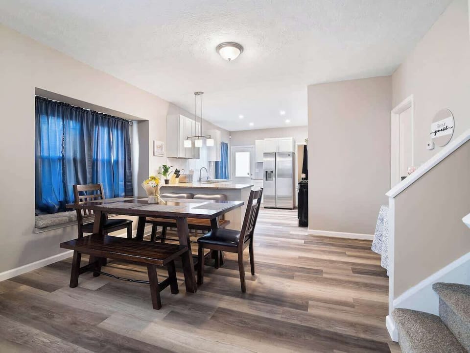 Cozy open space with leather sofas, wooden coffee table, and adjacent dining bench and chairs by bay window. Vinyl plank floors and blue curtains add warmth and style—perfect for relaxing and entertaining! ️️