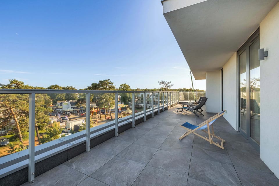 A long, open balcony bordered by glass railings stretches out toward a view of lush green treetops. A wooden deck chair sits ready for relaxation.