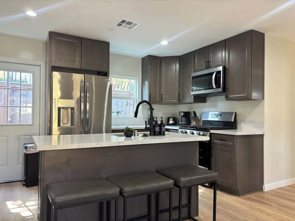 Modern kitchen with sleek dark cabinetry, stainless steel appliances, and a spacious quartz countertop with high top seating. Features a gas stove, large fridge, microwave, and black pull-down faucet. Bright, inviting, and perfect for group meals.