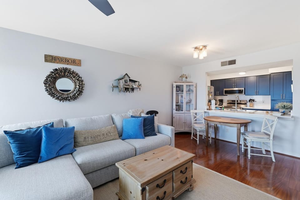 Living area featuring a gray sectional with blue pillows, wood coffee table, and hardwood floors leading to the kitchen.