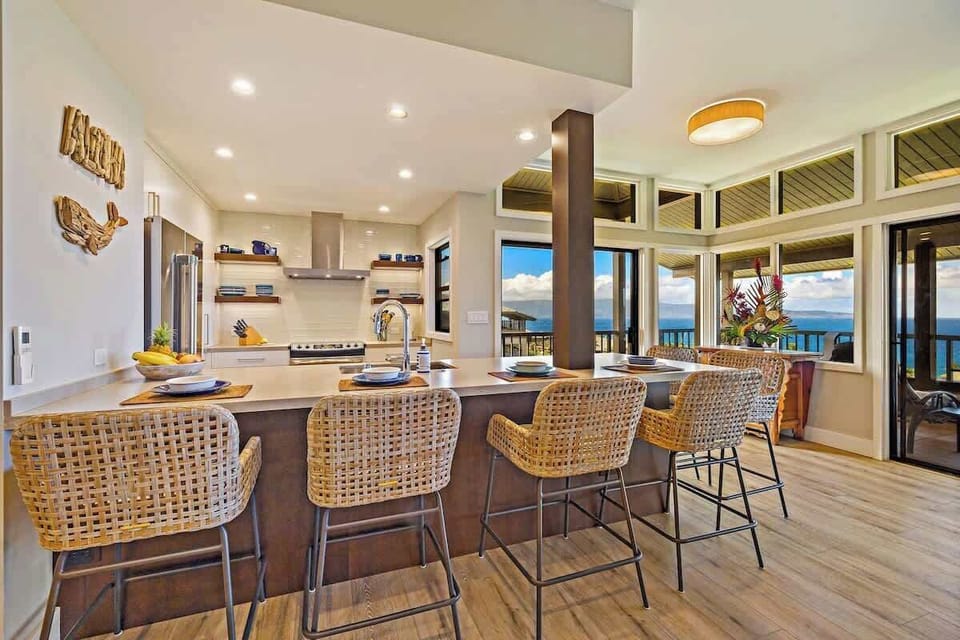 Modern open kitchen with spacious island, sleek appliances, and rattan barstool seating. Perfect for preparing home cooked meals, this bright space offers seating for the whole family while enjoying stunning Maui ocean views through large windows!