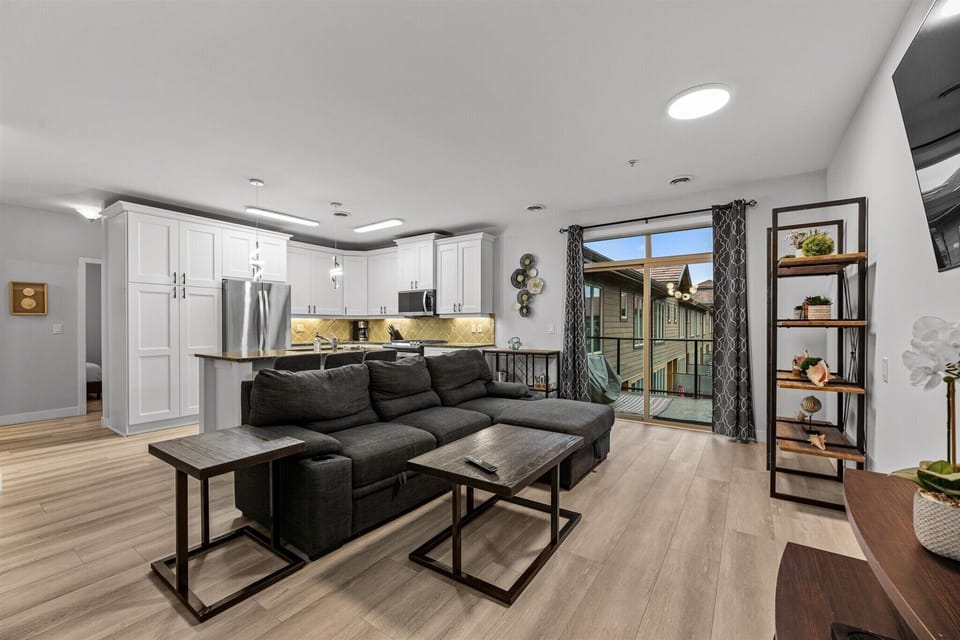 Bright and open living space featuring a cozy sectional sofa, smart TV, and sliding doors to a private balcony. The modern open-concept layout flows seamlessly into the fully equipped kitchen, perfect for relaxing or entertaining.
