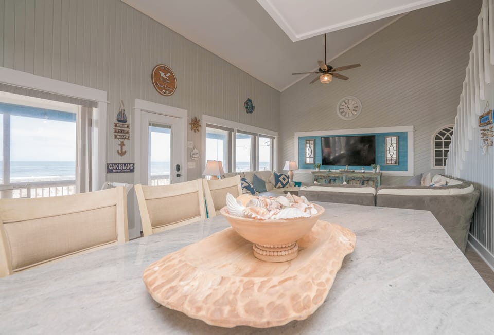 Bright dining area with table for six, distressed beach-style cabinet, bar seating, chandelier, and large windows overlooking the covered deck and Atlantic view.