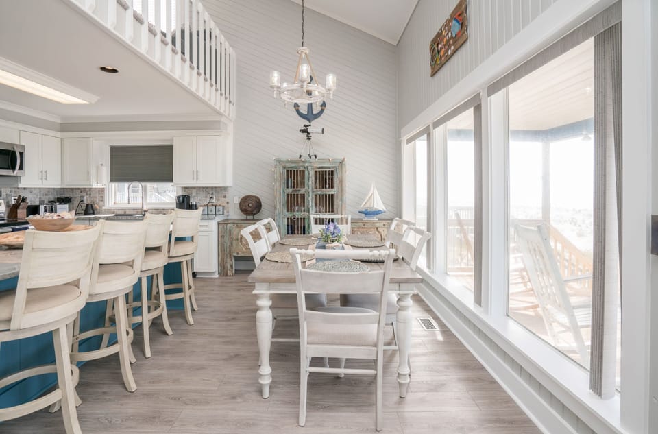 Bright dining area with table for six, distressed beach-style cabinet, bar seating, chandelier, and large windows overlooking the covered deck and Atlantic view.