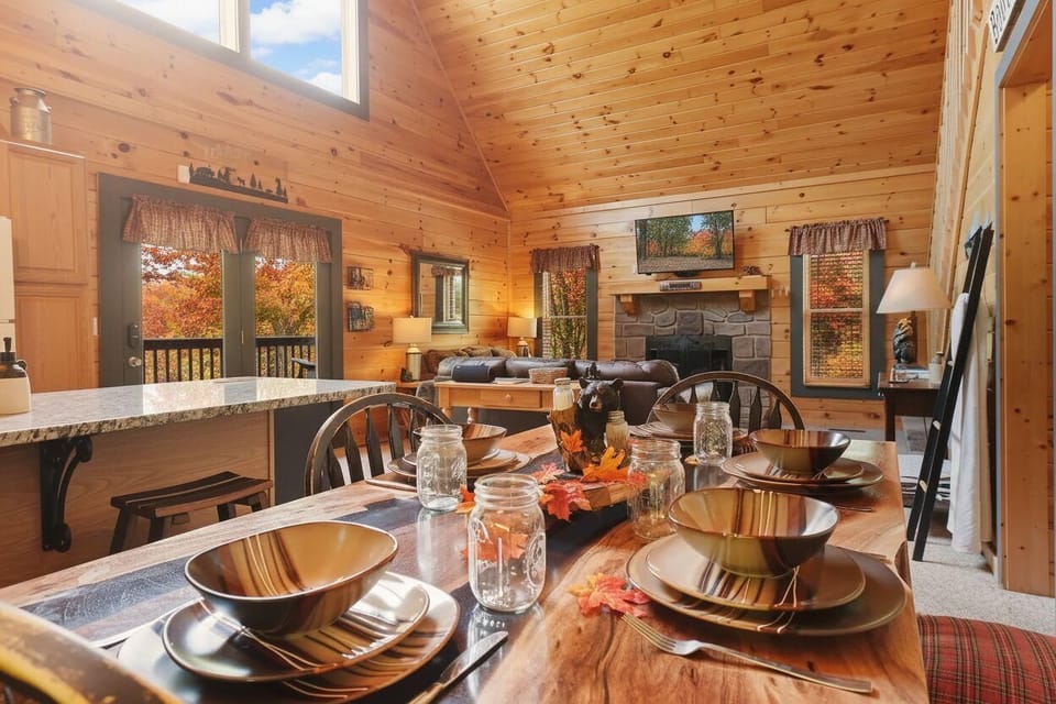Rustic dining area with a view of the living room and stone fireplace in a wood-paneled cabin.