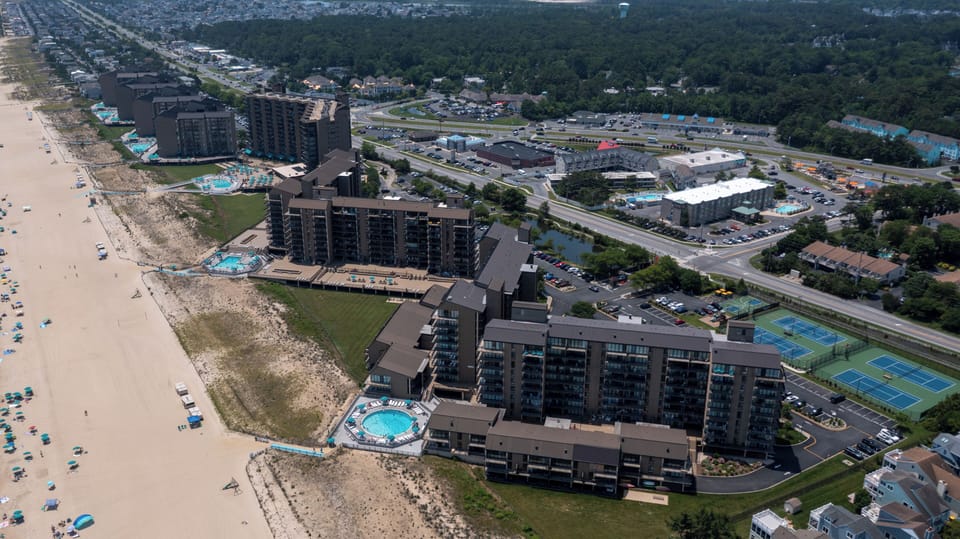 806 Harbor Sea Colony East Outdoor Swimming Pool with Ocean View - A ground-level perspective of an outdoor resort pool bounded by a glass safety fence overlooking the sandy beach and ocean coastline.