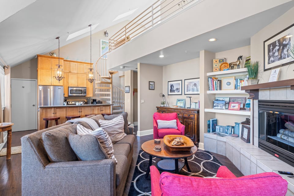 A cozy and eclectic living room with an open kitchen and loft with a spiral staircase, decorated with vibrant pink armchairs.