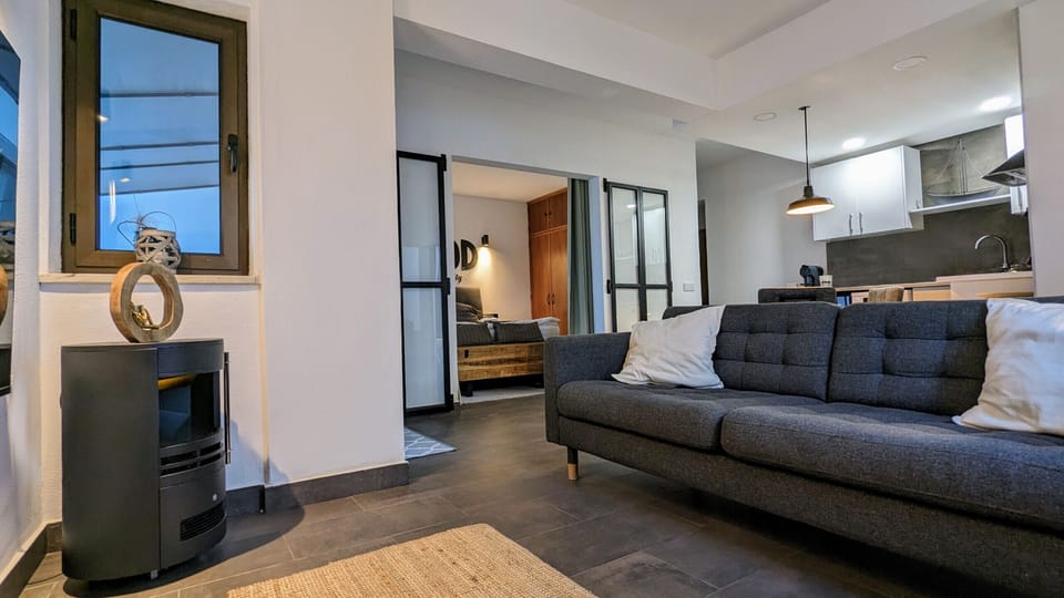 This bright and inviting living area welcomes you with a comfortable gray sofa, perfect for relaxing. A modern wood-burning stove, positioned next to a window that lets in natural light, provides cozy warmth on cooler evenings. The open-plan layout flows seamlessly into the kitchen area, equipped with white cabinetry and modern appliances – ideal for preparing meals while staying connected with your travel companions. An elegant glass partition with black frames allows you to separate the living area from the bedroom without sacrificing the feeling of openness. The bedroom itself is visible in the background, offering a tranquil retreat. The combination of dark tile floors and light walls creates a modern yet inviting atmosphere, perfect for relaxing and enjoying your stay in Lagos.