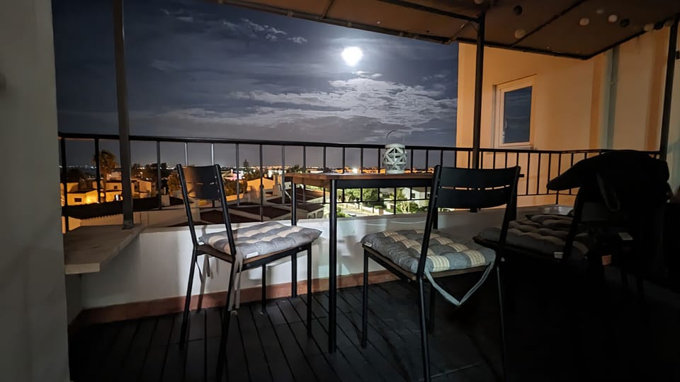 Experience the magical atmosphere of this balcony in Lagos at night. The full moon shines brightly in the sky, bathing the entire scene in a soft, mystical light. On the balcony itself, you'll find a table and chairs, inviting you to sit under the starry sky and enjoy the tranquility of the evening. Your gaze sweeps across the rooftops of the city, its lights twinkling in the distance, enlivening the night sky of Lagos. The light cloud cover around the moon enhances the dramatic yet peaceful ambiance. Imagine yourself ending your day here, perhaps with a glass of wine, surrounded by the stillness of the night and the enchanting moonlight.