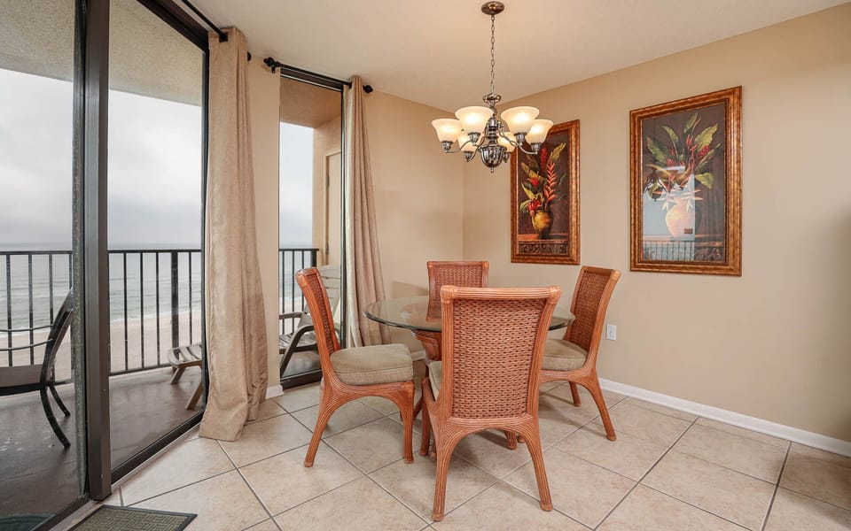 A round glass dining table surrounded by rattan chairs offers a cozy space with beach views through large sliding doors