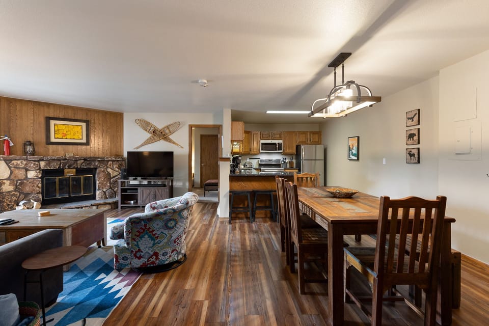 A cozy living and dining area with wood flooring, stone fireplace, TV, colorful rug, patterned armchair, wooden dining table with chairs, and a kitchen with stainless steel appliances in the background.