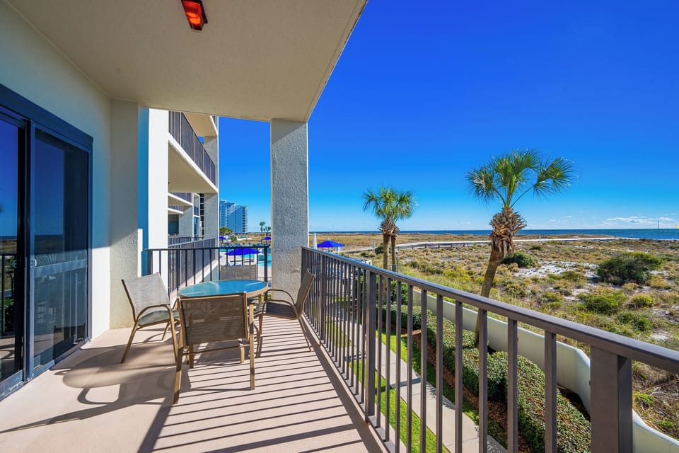 Balcony with a glass table and chairs overlooks lush greenery and the beach, framed by palm trees against a clear blue sky