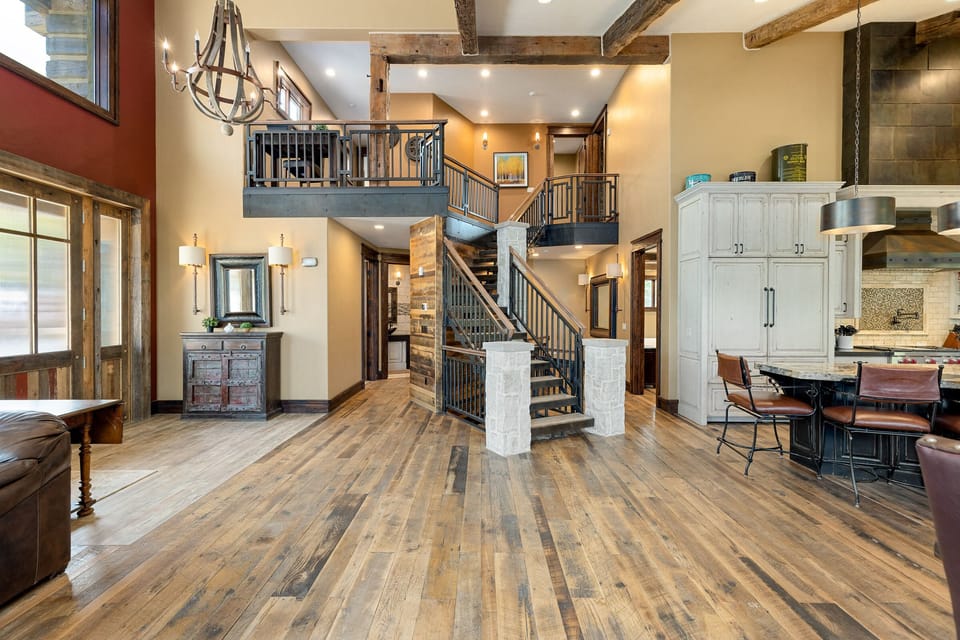 Spacious, rustic-style living area with wooden flooring, a central staircase, a chandelier, large windows, and an open kitchen with a breakfast bar and high chairs.
