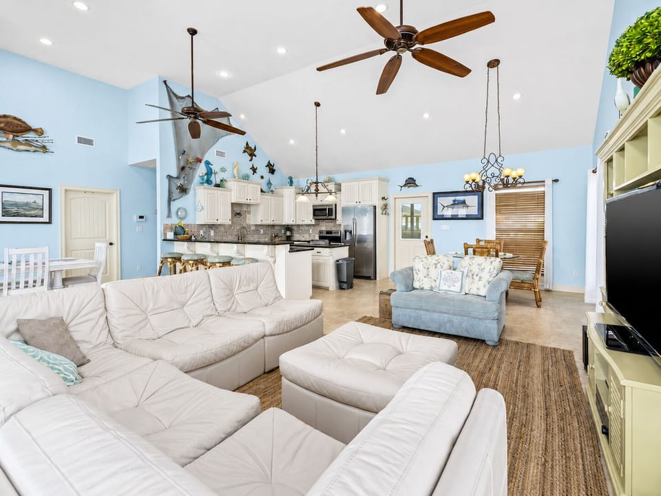 Open-concept living room with plush sectional seating, vaulted ceilings, ceiling fans, coastal décor, and direct views toward the kitchen.