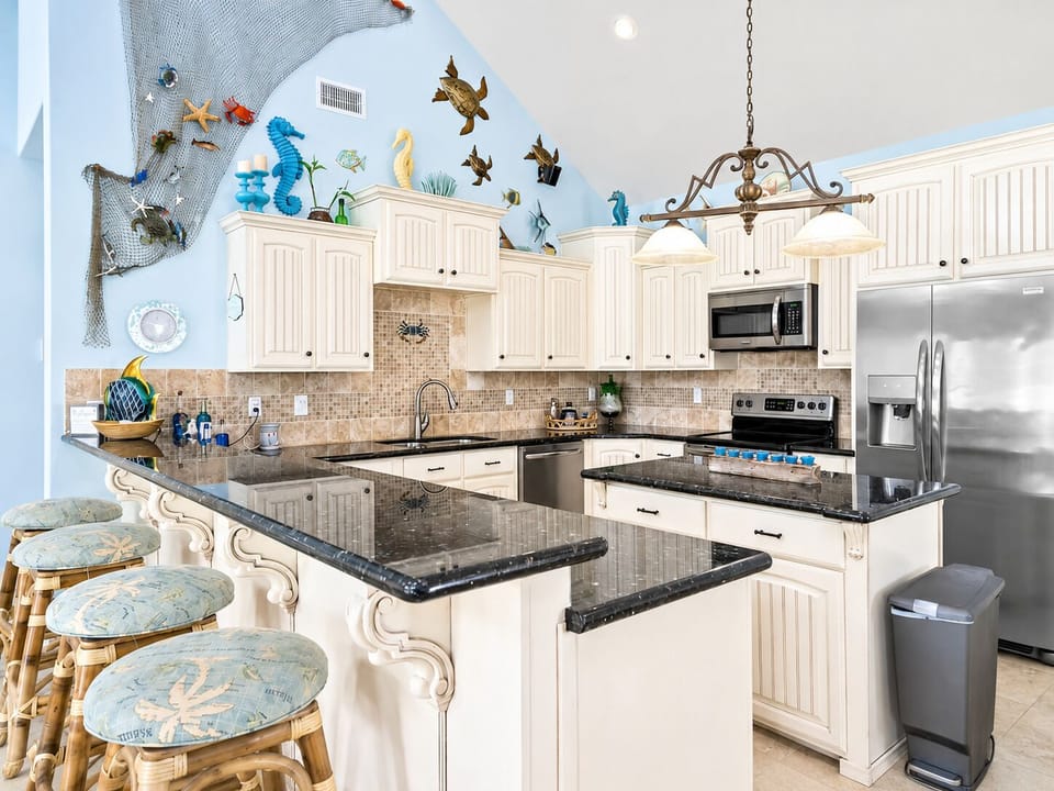 Fully equipped coastal kitchen with granite countertops, breakfast bar seating, white cabinets, stainless steel appliances, and generous workspace.
