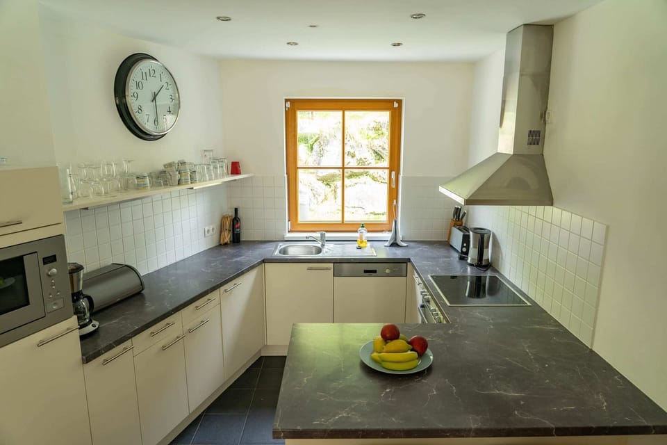 Sunlight bathes this contemporary kitchen, highlighting the sleek countertops and stainless steel appliances. A large window showcases a verdant backdrop, creating a serene ambiance for enjoying morning coffee or evening cocktails.