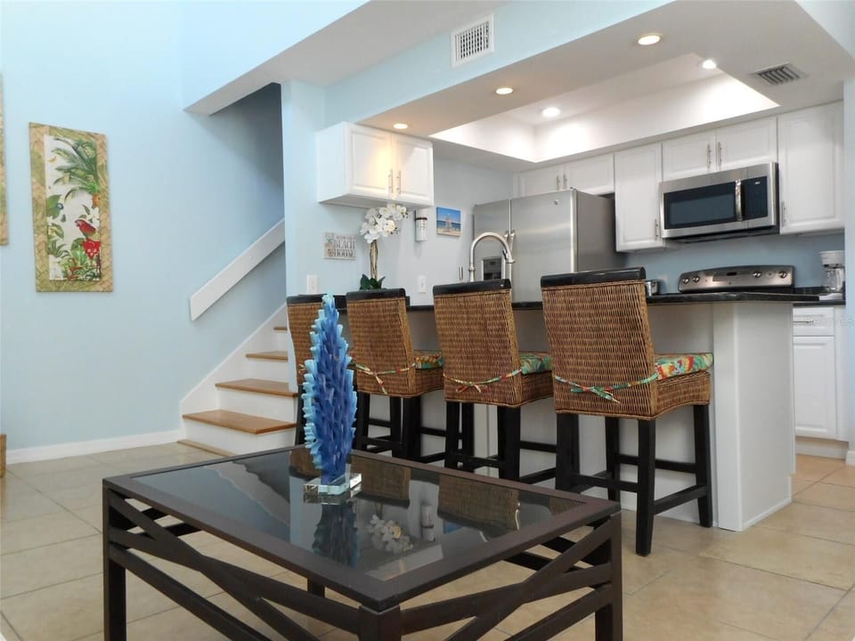 Kitchen bar with dark granite island, wicker bar stools, stainless steel appliances, and framed coastal artwork