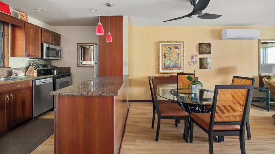 Gather around the glass dining table in your open-concept living space, where rich wood tones, tropical artwork, and an island kitchen blend comfort with island style for memorable meals together.