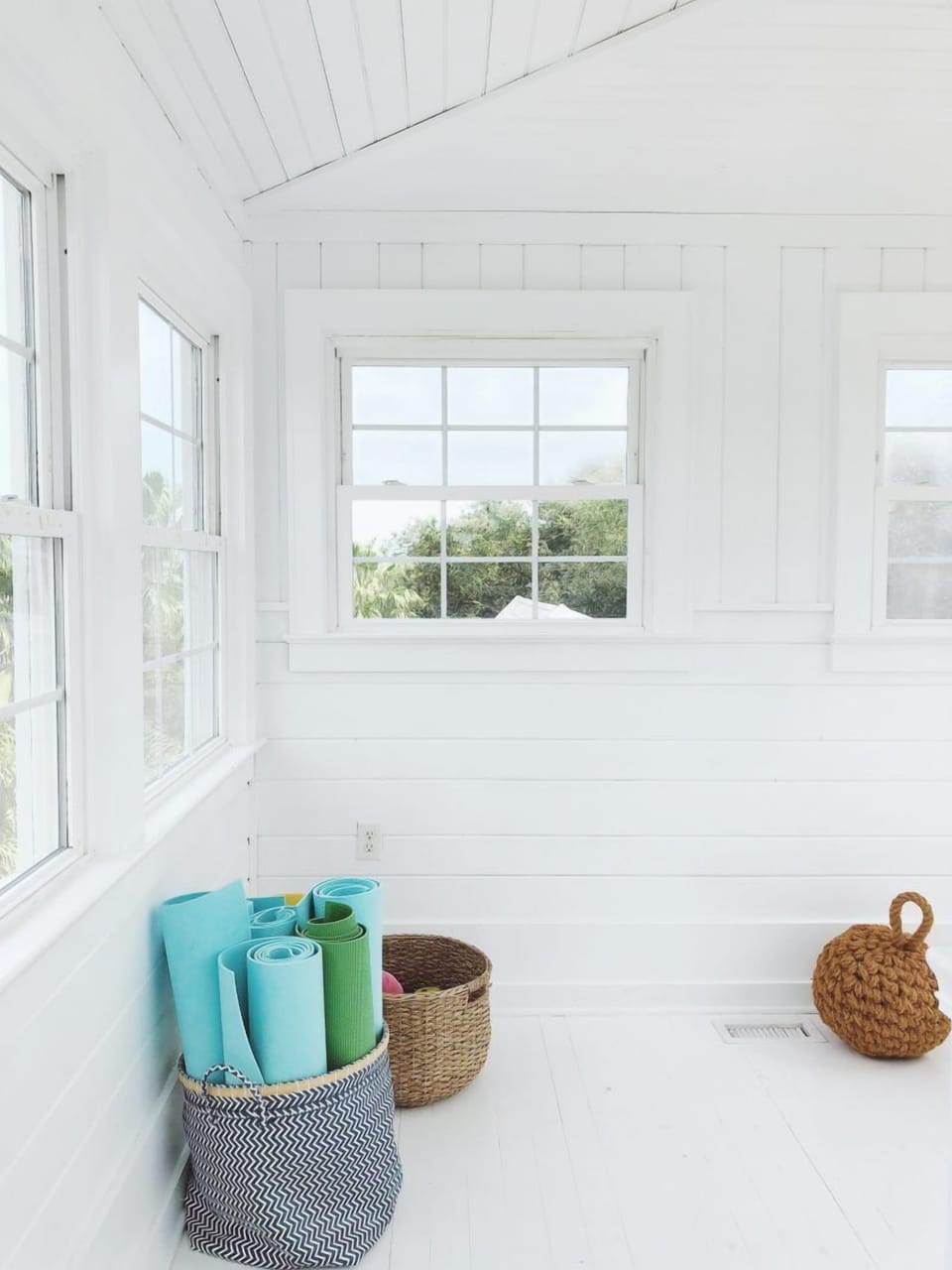 A bright white room with large windows. Yoga mats are stored in a basket on the floor, and a woven basket is nearby.