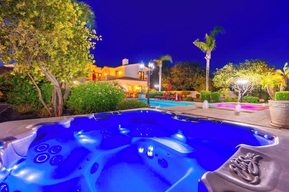 This beautifully lit evening scene showcases an outdoor hot tub area surrounded by lush tropical landscaping, illuminated poolside lounging zones, and a multi-level vacation villa with warm ambient lighting under a vibrant twilight sky.