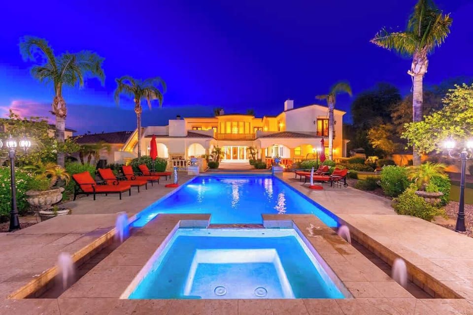 Stunning Mediterranean-style estate illuminated under a twilight sky, featuring an inviting pool with integrated spa, surrounded by swaying palms and vibrant landscaping.