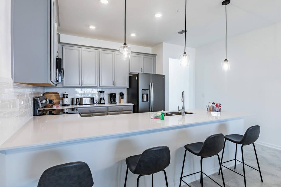 Experience a culinary delight in our stylish kitchen featuring modern gray cabinetry, a spacious quartz island, and chic black bar stools. The bright, energy-efficient lighting creates a vibrant atmosphere for your stay.