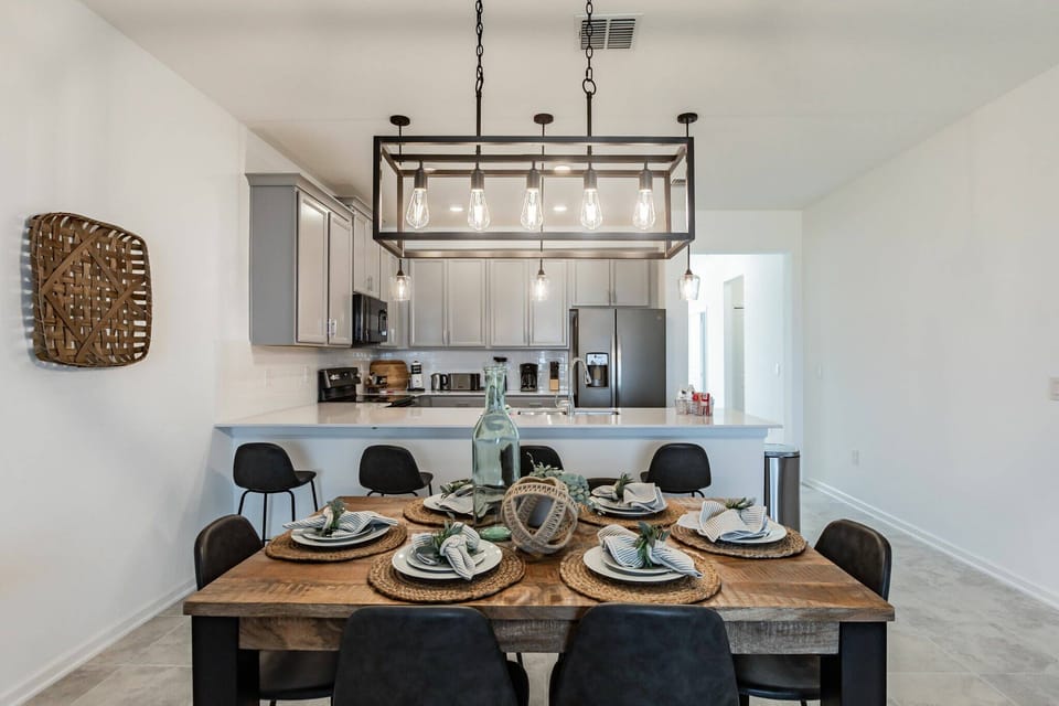Join us at the charming wooden dining table, elegantly set with stylish dishware, and soak in the warm ambiance from the stunning pendant light above while the modern kitchen in the background enhances your dining experience.