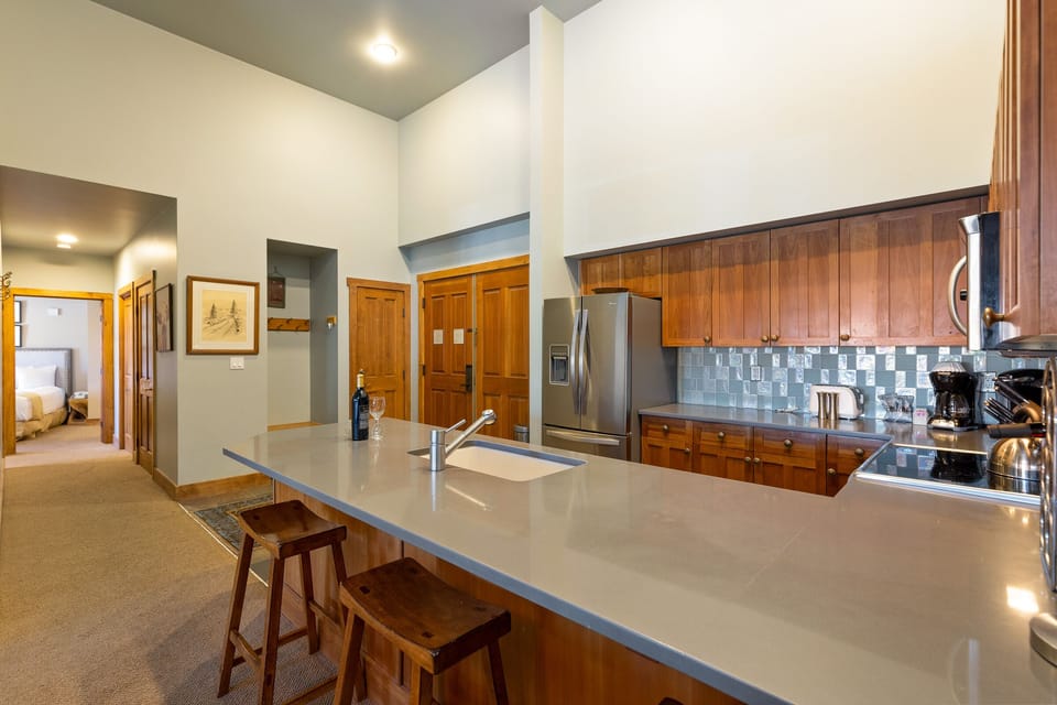 A spacious kitchen features wooden cabinets, a stainless steel refrigerator, bar stools at an island counter, and visible doorways to other parts of the house.