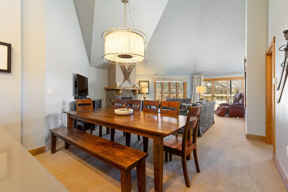 A dining room with a wooden table and bench seating, a chandelier overhead, and a view of a living room with armchairs and a large window showcasing an outside landscape.