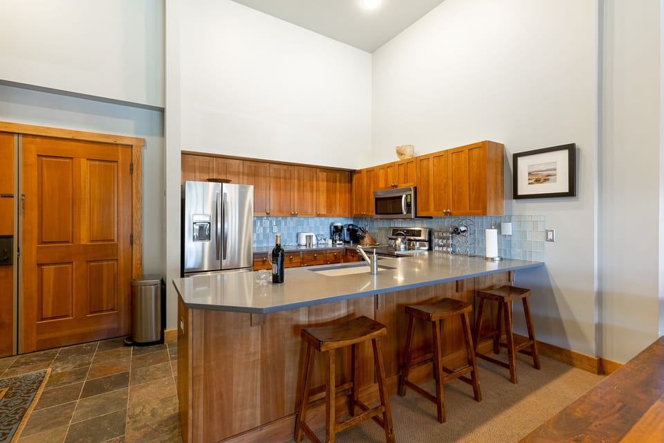 Modern kitchen with wooden cabinets, stainless steel appliances, a countertop with stools, and a framed picture on the wall.