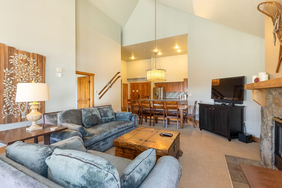 A cozy living room with a vaulted ceiling, gray couches, a wooden coffee table, a wall-mounted TV, dining table, and a stone fireplace. The room features rustic decor and a view into the kitchen area.