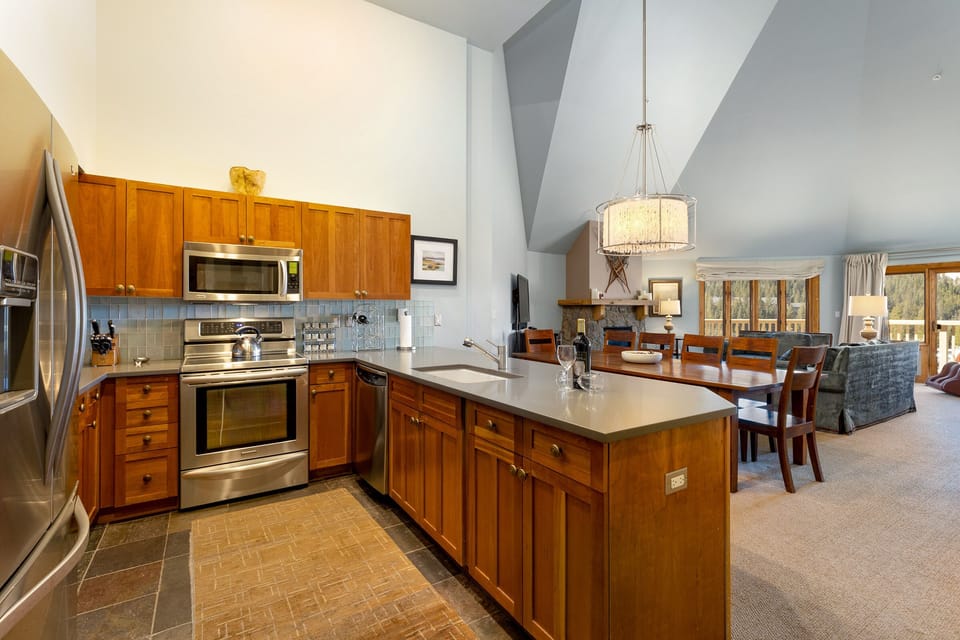 A modern kitchen with wooden cabinets, stainless steel appliances, and an island faces an open dining and living area with a high ceiling and large windows.
