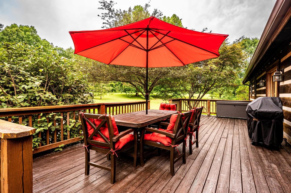Wood deck with dark wood dining table, red-cushioned chairs, matching umbrella, gas grill, and forest views.