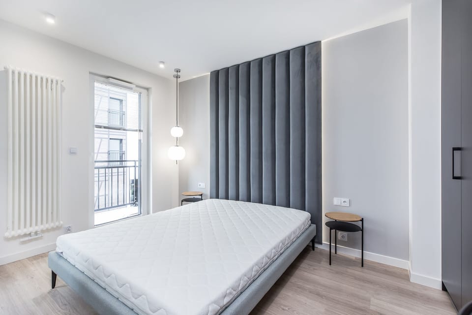 A minimalist bedroom with a large bed, soft neutral bedding, and ample natural light coming through large windows, creating a serene and peaceful space.