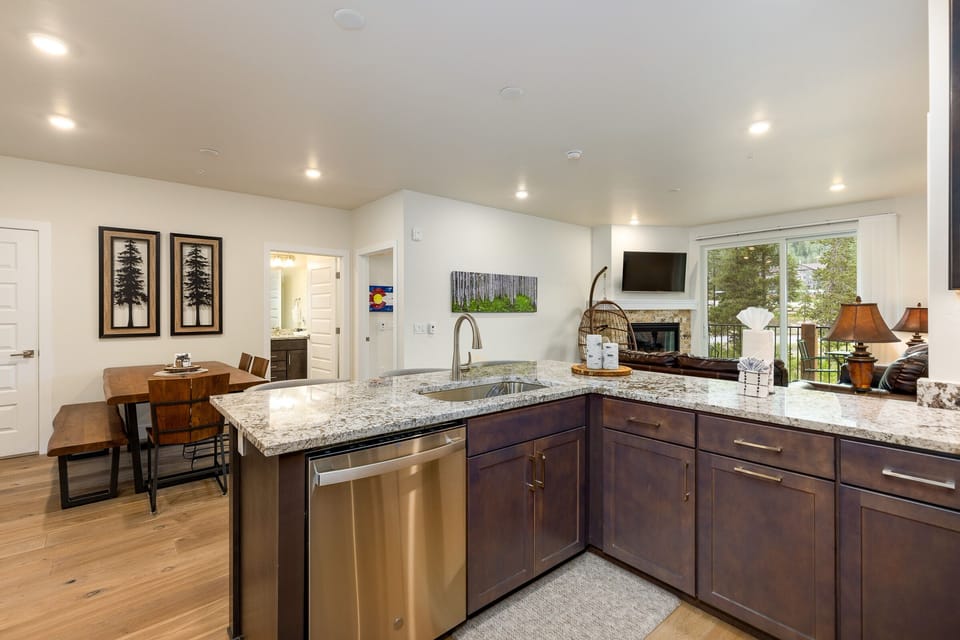 Modern open-concept kitchen with granite countertops, stainless steel appliances, and wood cabinets, leading into a living area with a fireplace and a view of an outdoor patio.