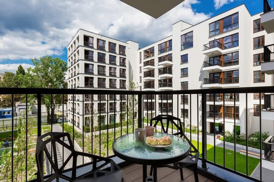 A balcony with an elegant black round table and two chairs, overlooking a modern residential complex surrounded by greenery.