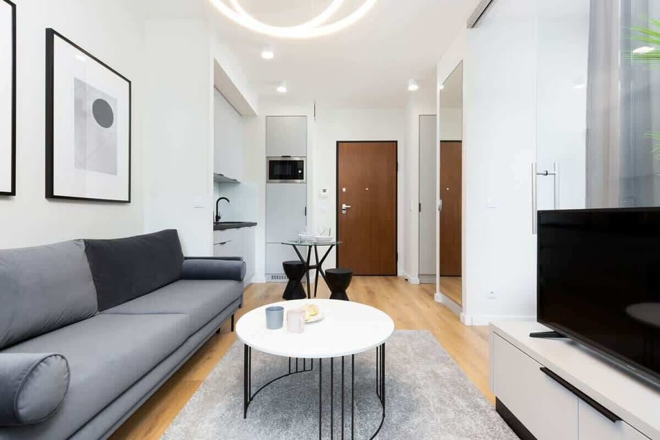 A spacious living room featuring a modern gray sofa, a white coffee table, and minimalist framed pictures on the wall. Bright colors and a large window add a sense of openness.