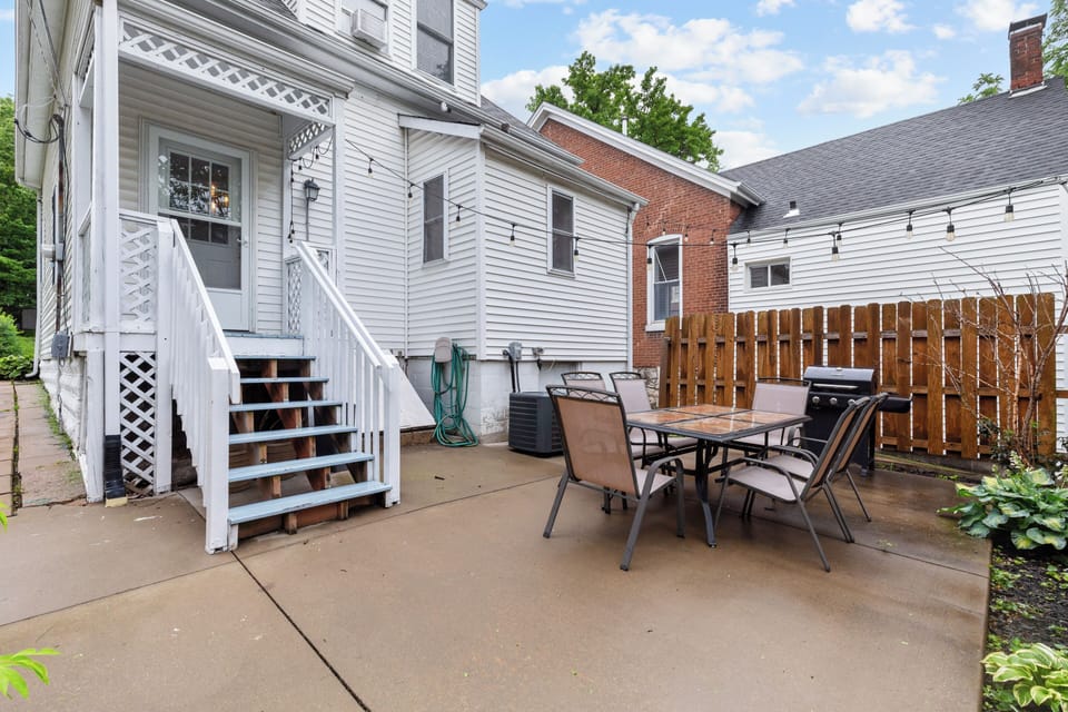 Rear-house view with raised back entrance, patio dining set, grill, string lights, and planting beds.