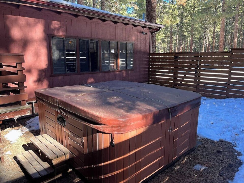 Hot tub area with hot tub cover, steps, privacy fence, house exterior, windows, snow, and trees.