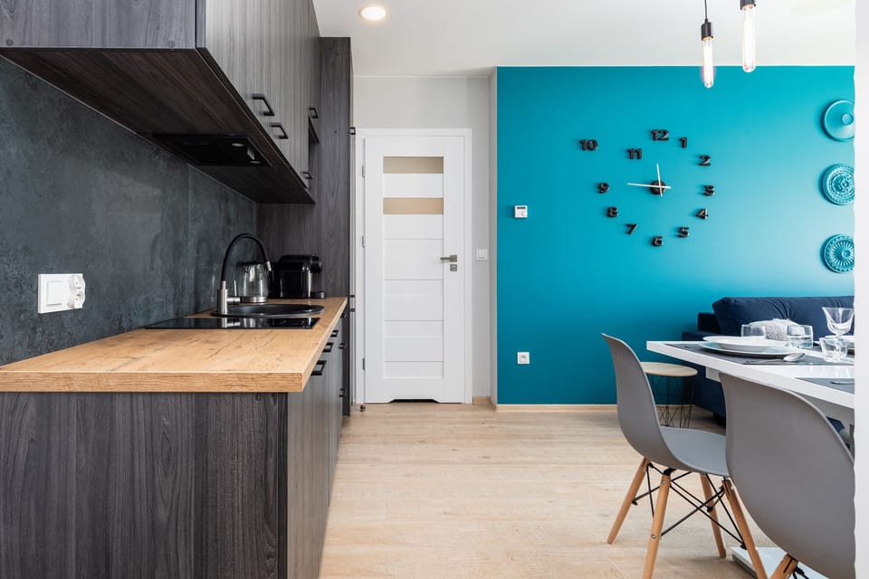 A kitchen with gray wooden cabinets, black countertops, and stainless steel fixtures. The turquoise wall clock adds color to the space.