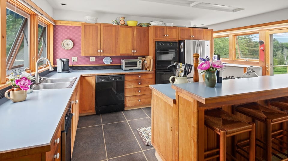 A spacious kitchen with wooden cabinets, black appliances, a center island with stools, and a window view of trees and mountains. Flower vases are placed on the counters.