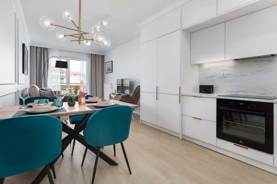 A modern open-plan kitchen and dining area with white cabinetry, a marble backsplash, and a black oven, paired with teal dining chairs for a touch of color.

