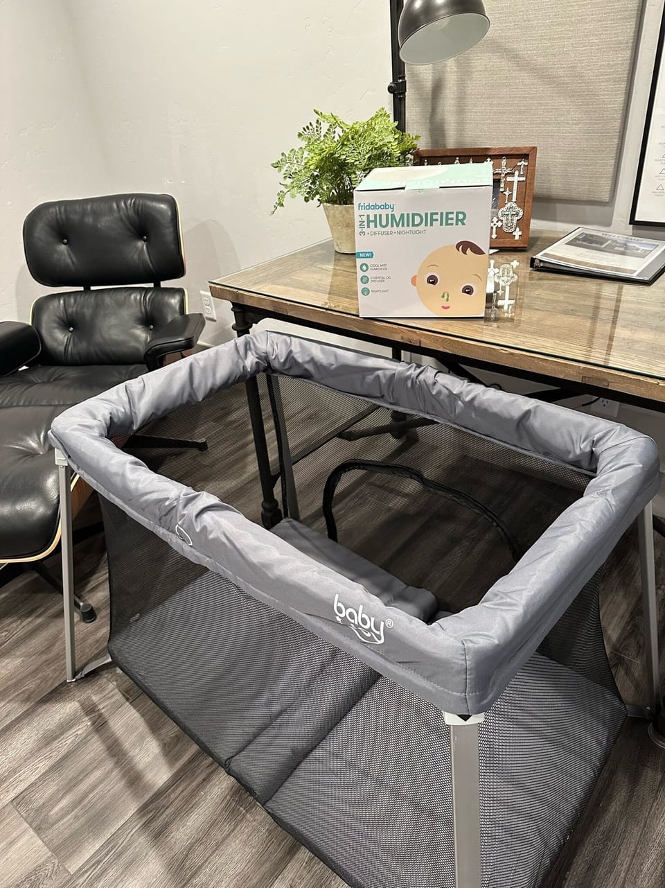A gray infant playpen is set up on a wood floor next to a desk with a black chair. On the desk, there is a baby humidifier box and a potted plant.