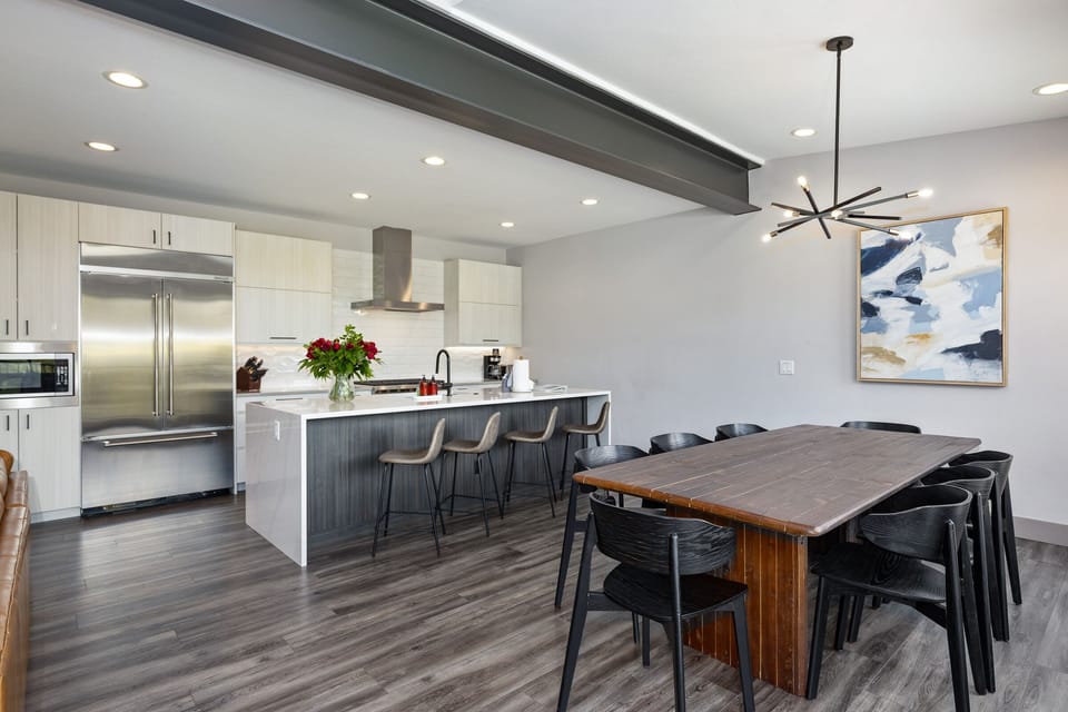 Modern kitchen and dining area with stainless steel appliances, a large island with bar stools, a wooden dining table with black chairs, and a contemporary pendant light.
