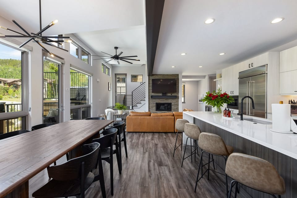 A modern, open-concept kitchen and living area with a large island, sleek appliances, a wooden dining table, leather couch, and a fireplace. Large windows provide natural light and views of greenery.