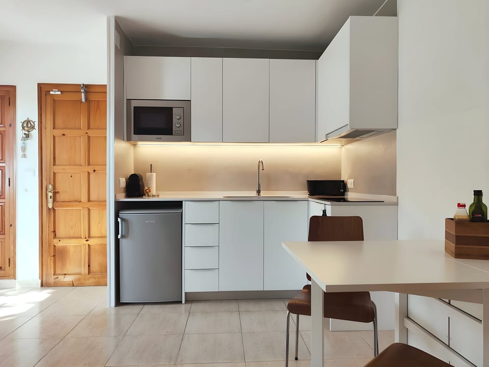 The bright and modern kitchen boasts sleek white cabinets and a compact layout. A small refrigerator, microwave, and coffee machine offer convenience for preparing meals, while a dining table and chairs provide a comfortable space for enjoying them. Natur