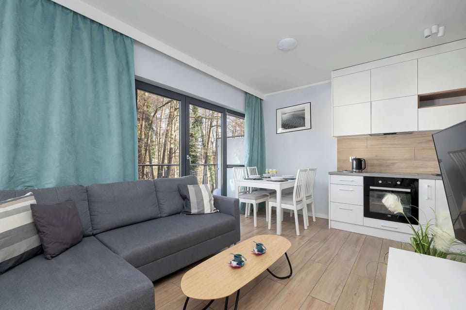 A living area with a gray sofa placed near large windows. Curtains frame the windows, and natural light fills the room. The seating area looks cozy and ideal for relaxing.