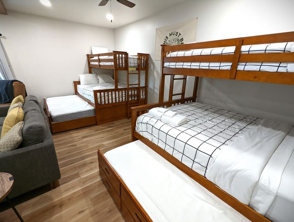 Your spacious bunk room sleeps the whole crew in cozy wooden beds with crisp linens, warmed by a ceiling fan and brightened by sunlight streaming through clean white walls.