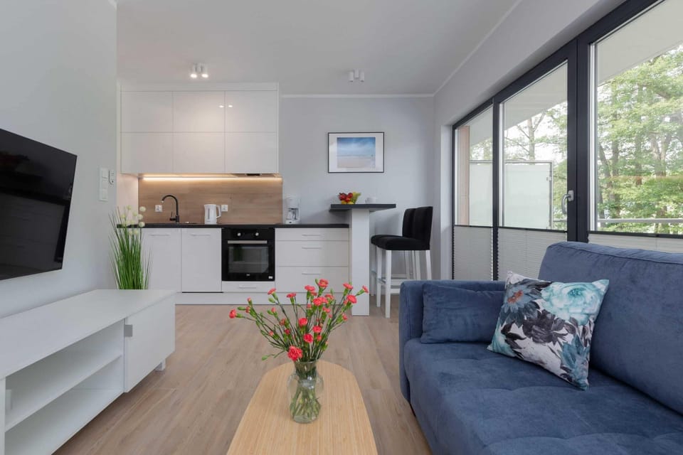 Another cozy living room contains a gray sofa, a wooden coffee table with flowers, a mounted TV, and large windows letting in sunlight.